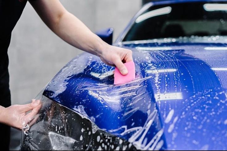 Technician carefully installing clear paint protection film on a car’s front bumper to shield the paint from scratches.